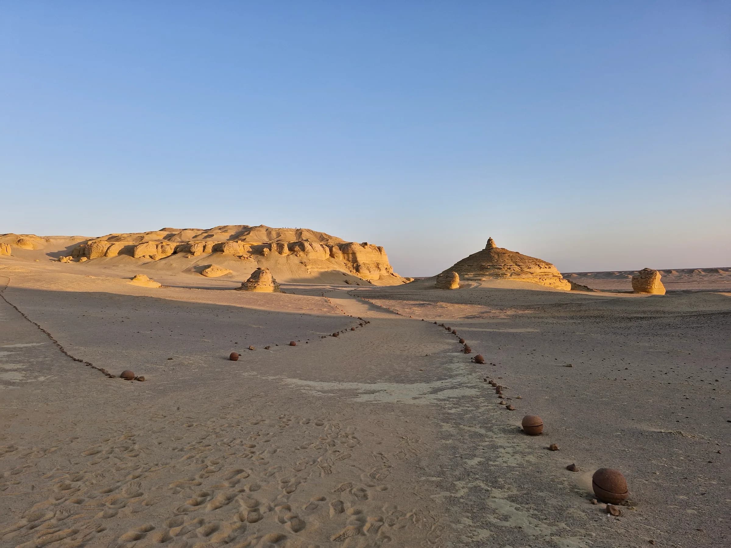 A desert landscape with a path of spherical stones leading towards unique sand-colored rock formations under a clear sky.